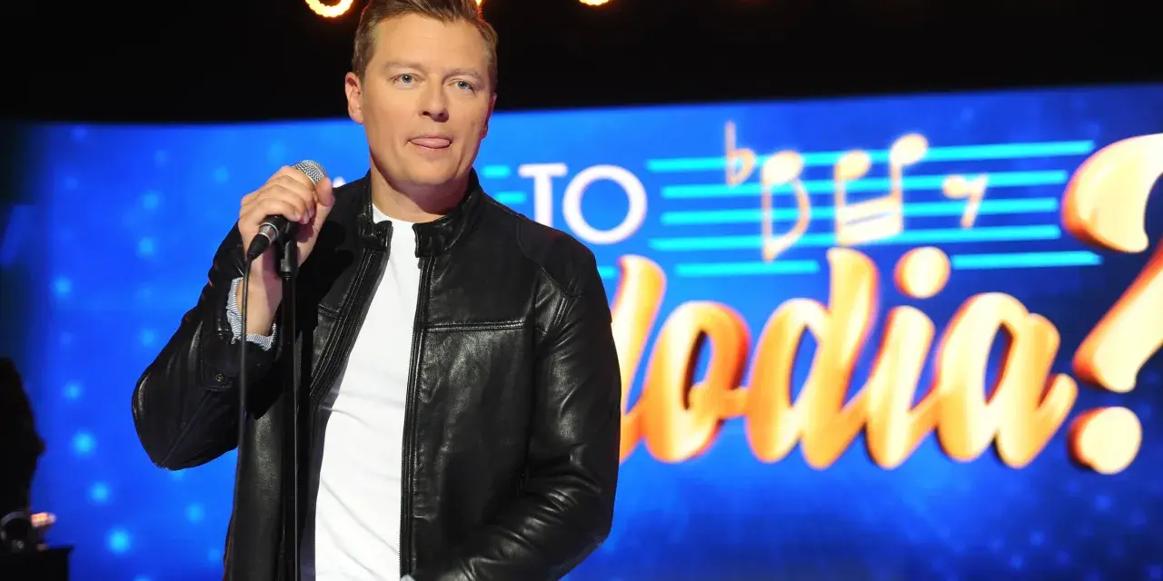 Rafał Brzozowski z mikrofonem na scenie, w tle napis "Jaka to melodia?".