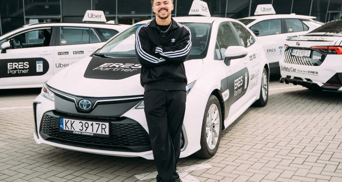 Młody mężczyzna w dresie stoi przed białymi samochodami Uber z napisem "ERES Partner". To jego praca prywatny kierowca.