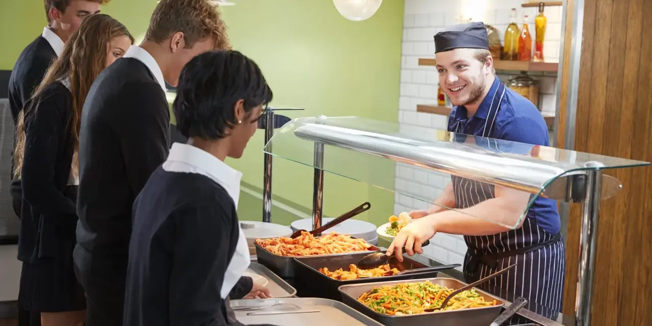 Des élèves font la queue pour le déjeuner à la cantine. Un employé souriant sert des légumes. Un contrat extra restauration est en cours de discussion.