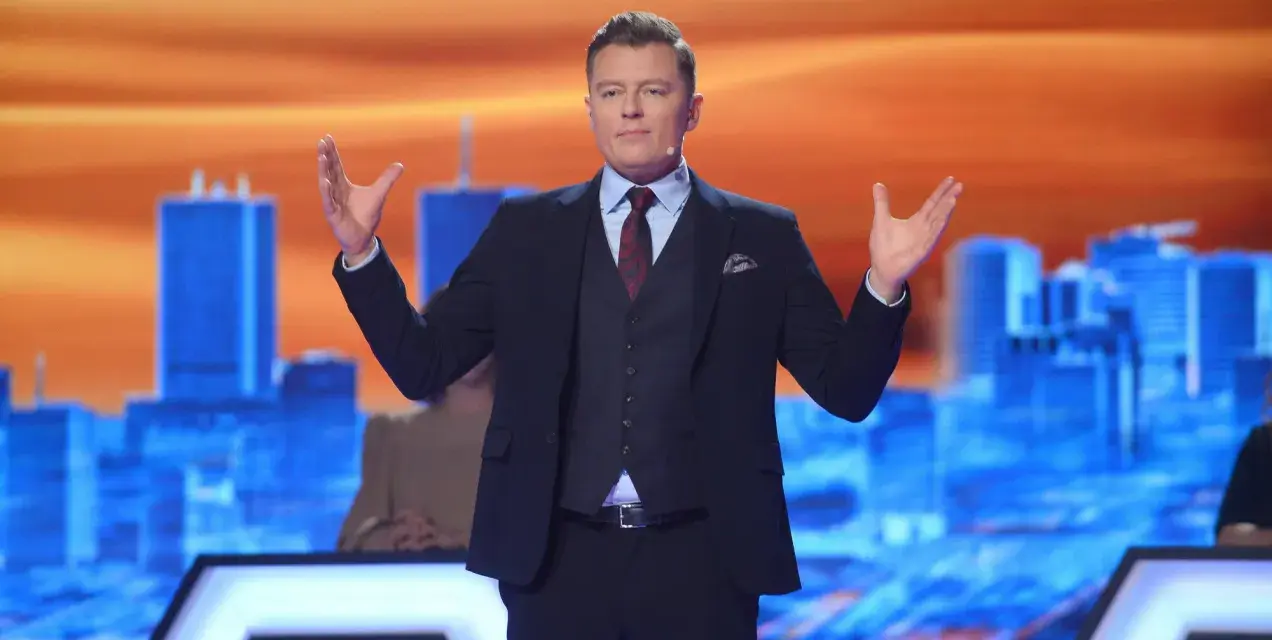 Rafał Brzozowski w garniturze, z rękami uniesionymi na boki, prowadzi program "Jaka to melodia?".