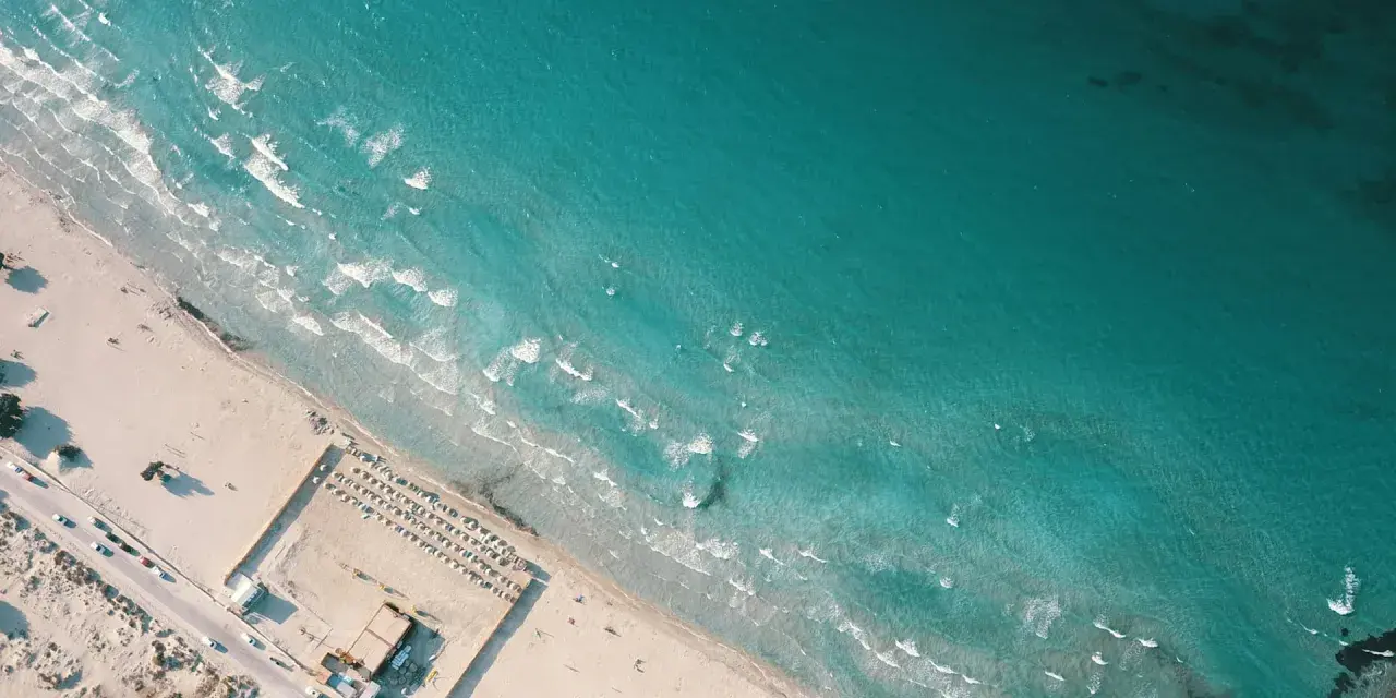 Izmir nad Morzem Egejskim. Piękna, piaszczysta plaża z turkusową wodą i falami. W oddali widać leżaki i parasole.