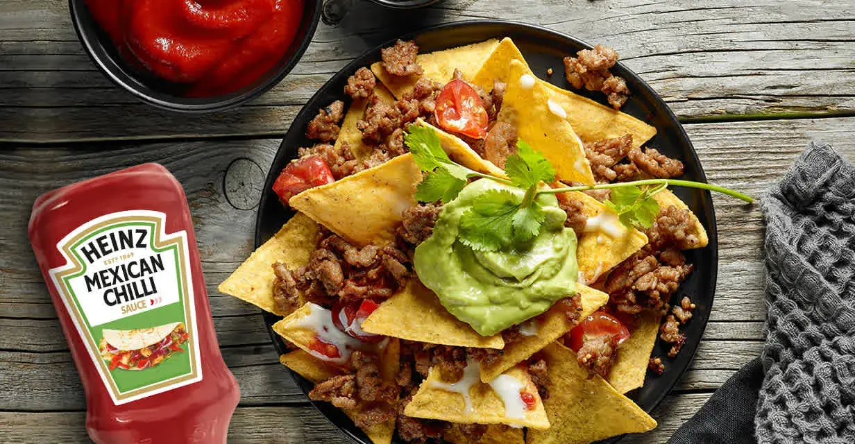Nachos con carne, guacamole y salsa en un plato negro, con botella de Heinz y cuencos de salsa.