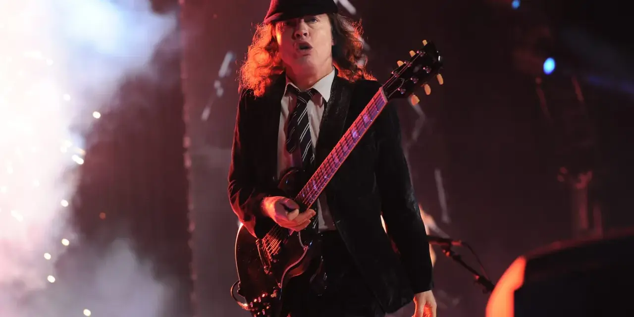 Ein Mann mit Hut und Schlagzeug spielt auf einer ACDC Gitarre, umgeben von Rauch und Lichtern.
