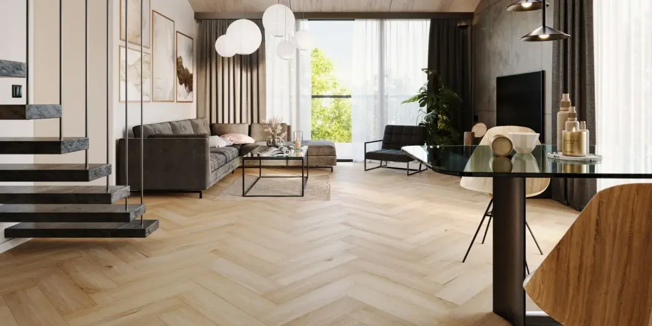 Nowoczesny salon z szarą sofą, szklanym stolikiem i jadalnią. Wzrok przyciągają panele bez dylatacji w progu, tworzące jednolitą, drewnianą podłogę w jodełkę.