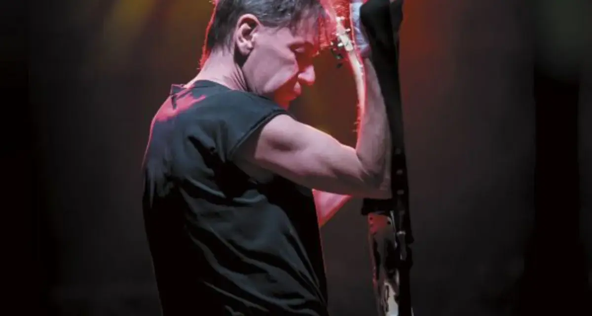 Muzyk z zespołu KSU z gitarą na scenie, w czarnej koszulce i z charakterystyczną fryzurą.