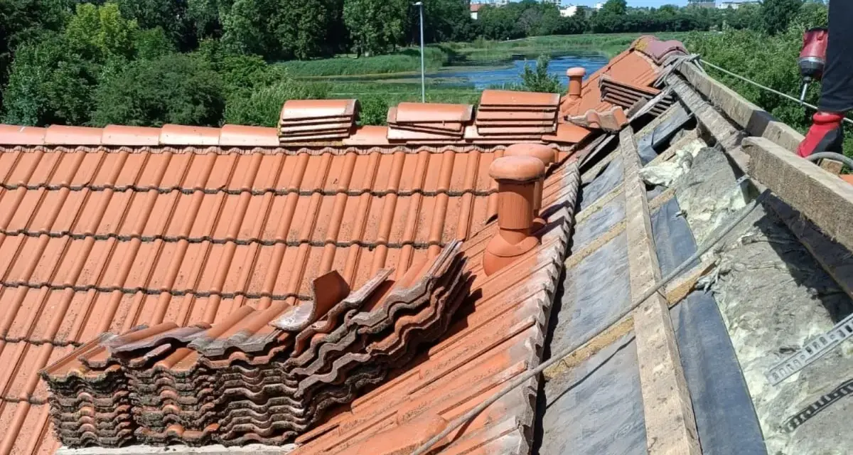Trwa wymiana dachu. Stare dachówki są ułożone w stosy, a pod nimi widać izolację i drewniane elementy konstrukcji.