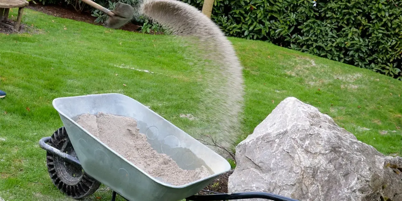 Schubkarre mit Sand, während eine Schaufel Sand auf den Rasen rieselt.