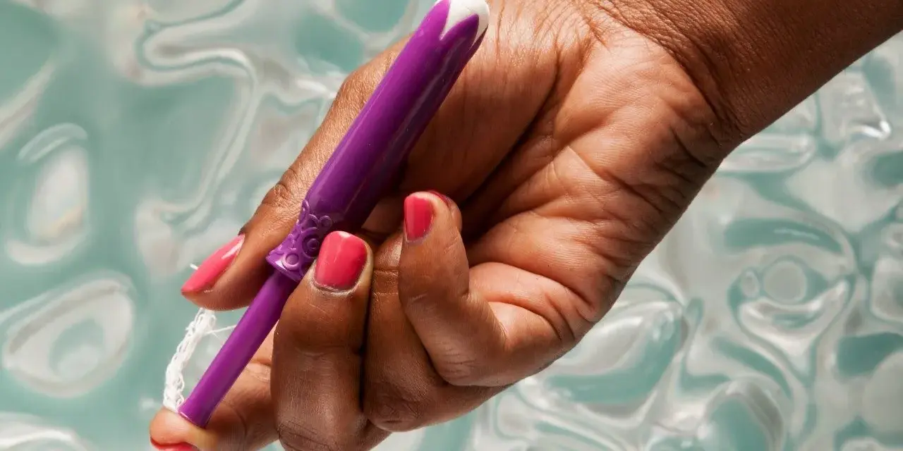 Dłoń z różowym lakierem trzyma fioletowy aplikator tamponu. Pokazano, jak zaaplikować tampon, trzymając go za sznurek.