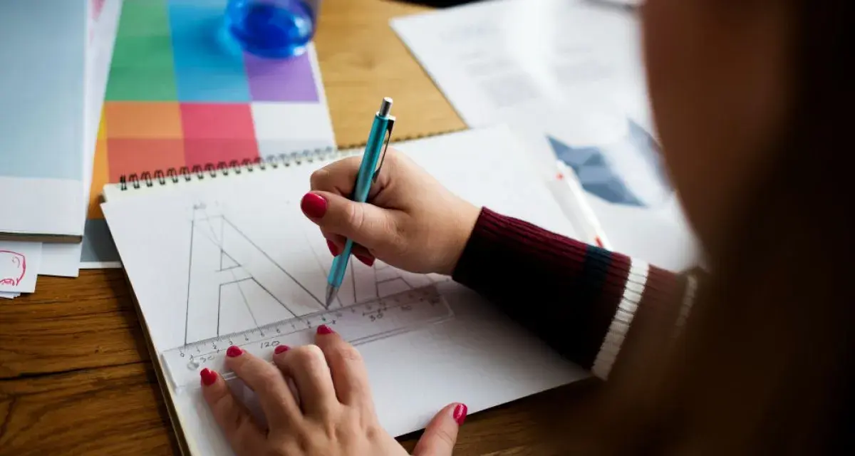 Ilustrator pracuje nad projektem, zamieniając litery na krzywe. Na stole leżą przybory do rysowania i próbnik kolorów.
