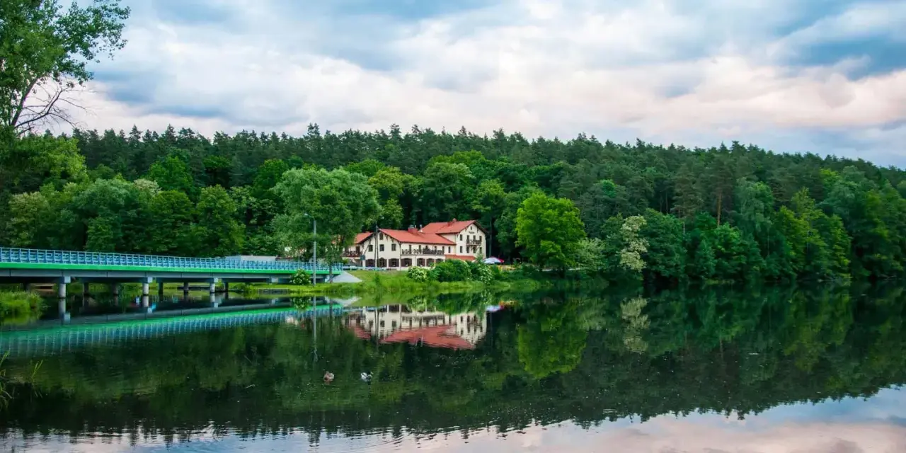 Malowniczy hotel nad jeziorem, z mostem i lasem w tle. Idealne miejsce na wypoczynek i odkrywanie tleń atrakcji.