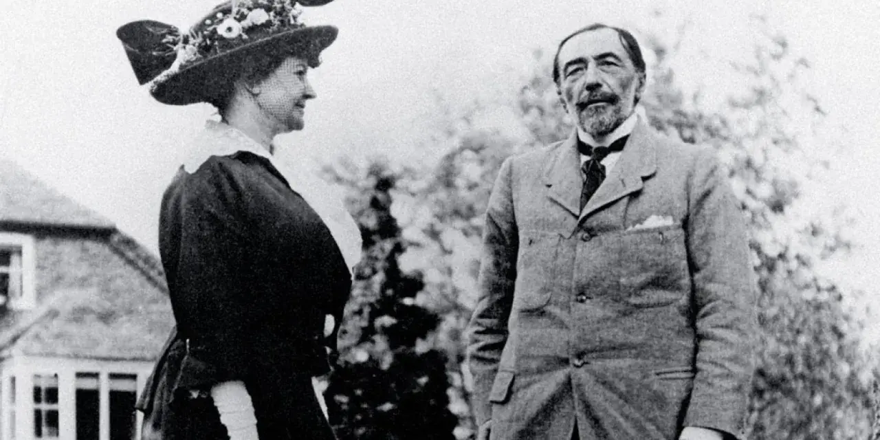 Józef Korzeniowski (aktor) w eleganckim garniturze stoi obok kobiety w ozdobnym kapeluszu.