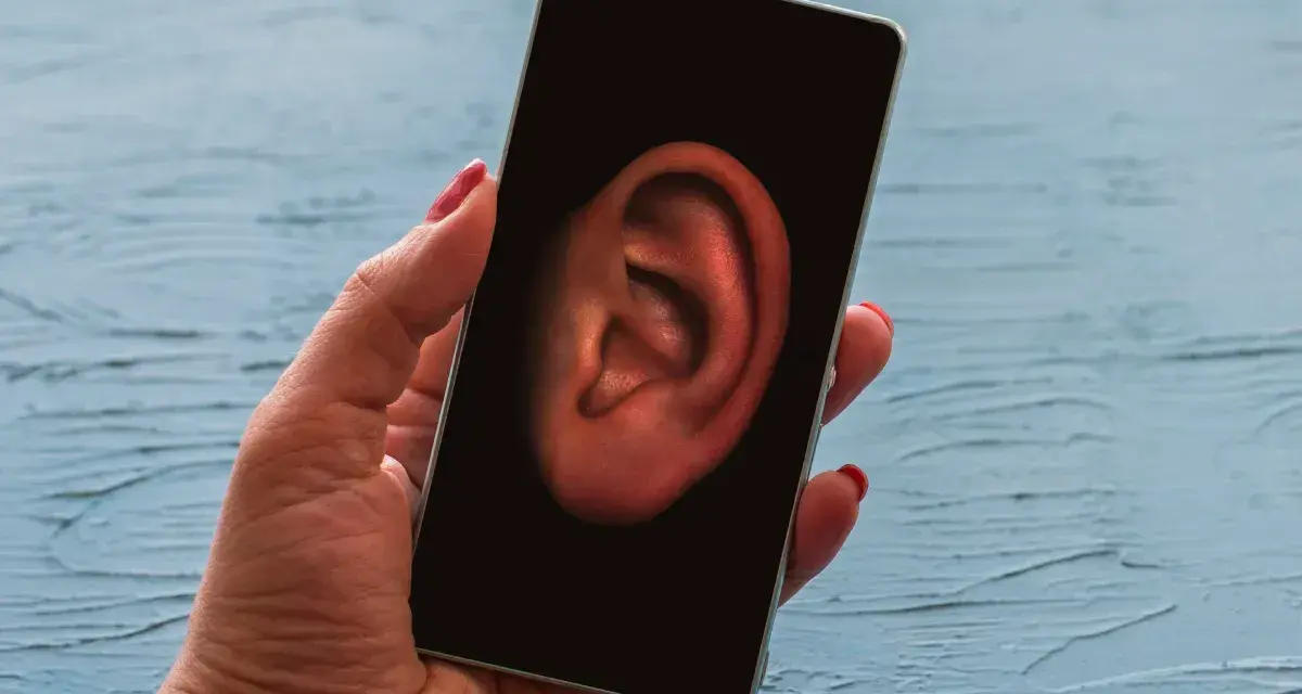 Dłoń trzyma smartfon z uchem na ekranie. Czy Twój telefon jest na podsłuchu? Sprawdź to!