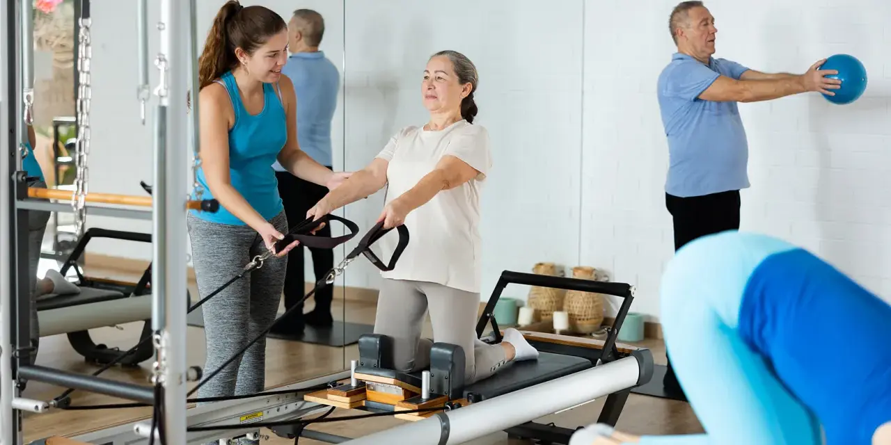 Frauen und Männer trainieren im **Reha-Sport** auf Pilatesgeräten. Eine Trainerin hilft einer älteren Frau, während ein Mann mit einem Ball übt.