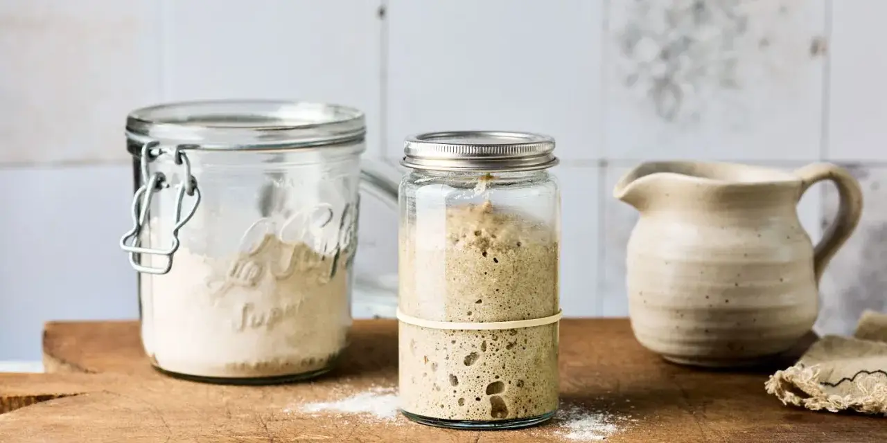 Zwei Gläser mit Sauerteig Weizen, eines mit aktivem, blasigem Sauerteig, das andere mit trockenem Ansatz. Daneben ein kleiner Krug.