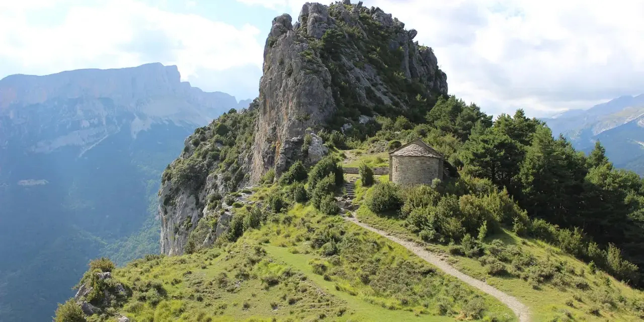 Pequeña ermita de piedra en la ruta de las ermitas, rodeada de vegetación y con imponentes montañas al fondo.