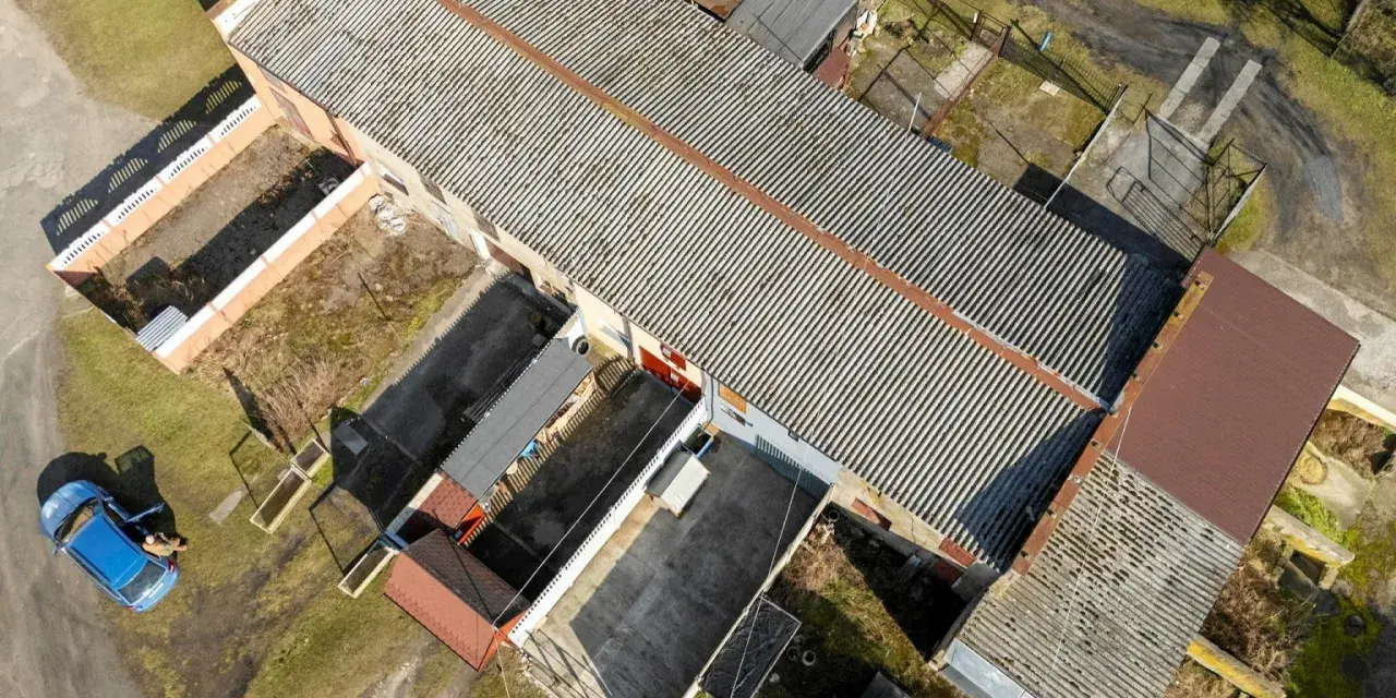 Długi budynek gospodarczy na zgłoszenie z falistym dachem, otoczony innymi mniejszymi obiektami i zielenią. Obok stoi niebieski samochód.