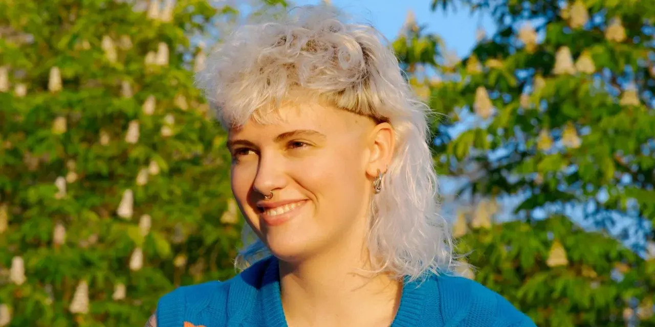 Persona sonriente con un moderno mullet rubio, aretes y un suéter azul con rayas naranjas, frente a un árbol en flor.