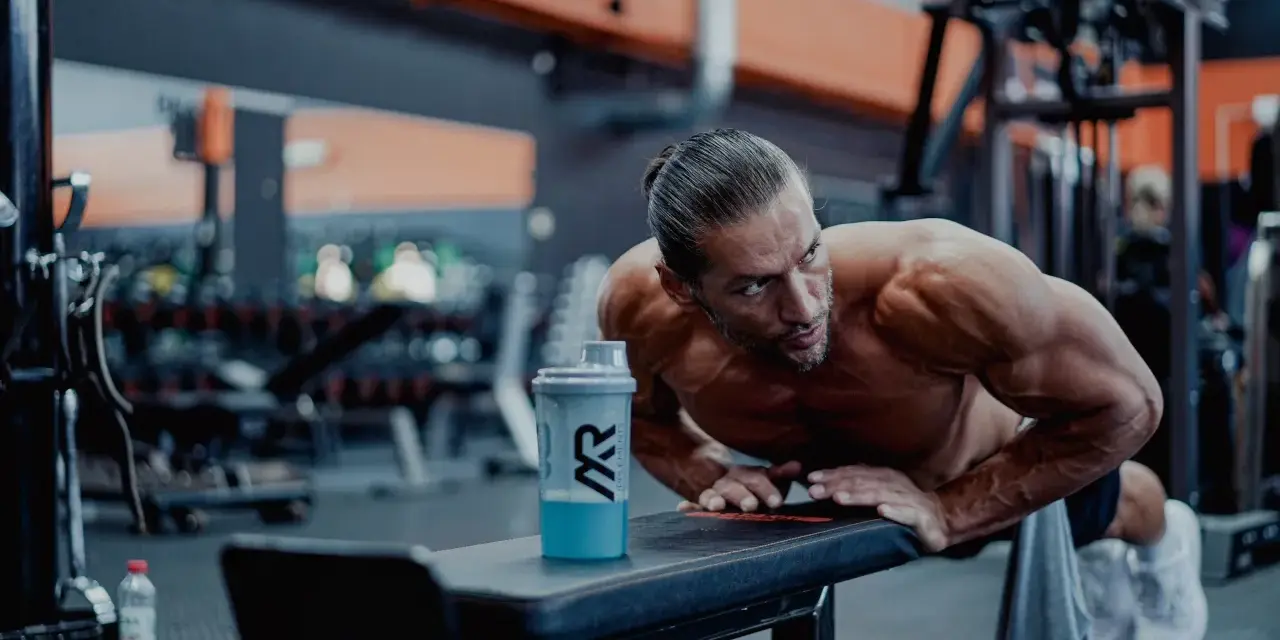 Man gör armhävningar på en bänk i gymmet, en shaker med blå dryck står bredvid. Detta är ett exempel på hur byggs muskler.