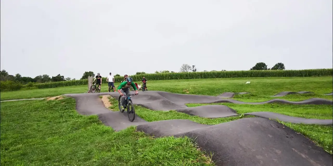 A group of people enjoy riding bikes on a winding pump track, with rolling hills and a cornfield in the background.
