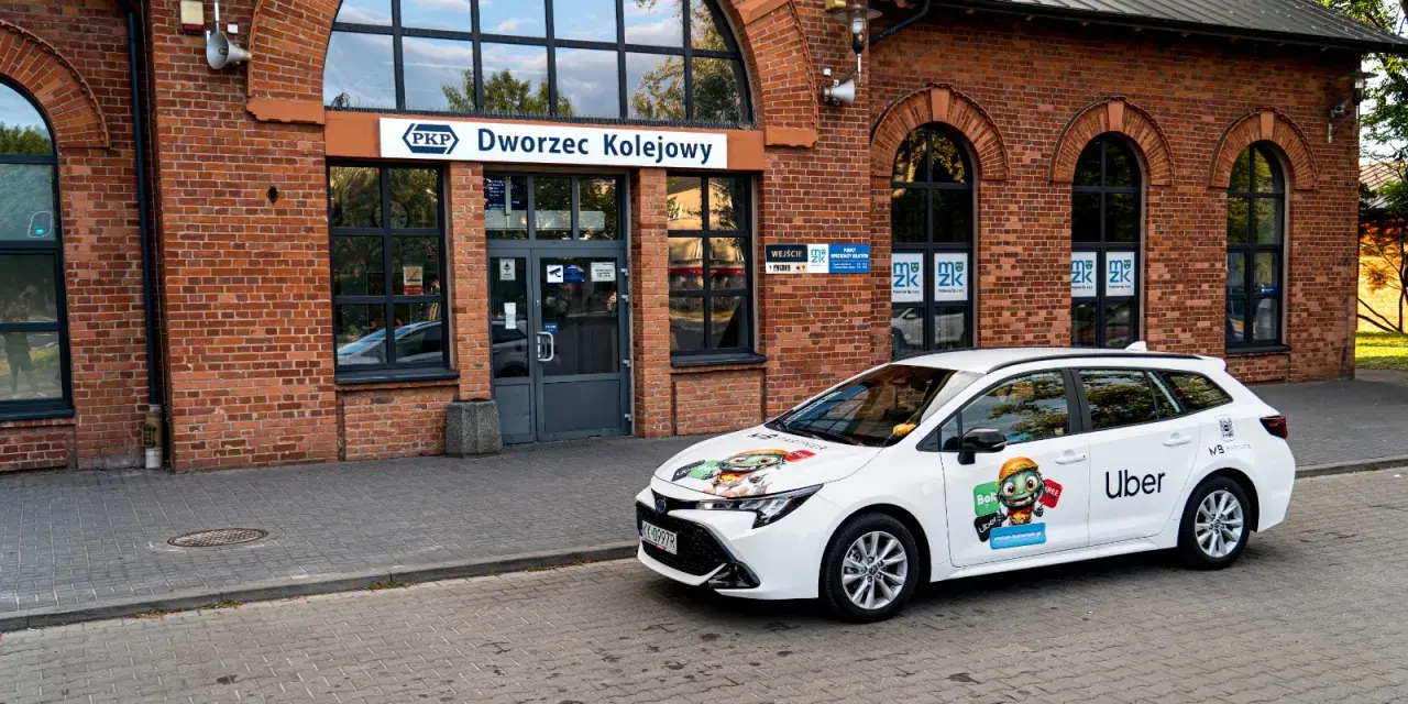 Biały samochód Ubera zaparkowany przed dworcem kolejowym. Kierowca z własnym samochodem czeka na pasażerów.