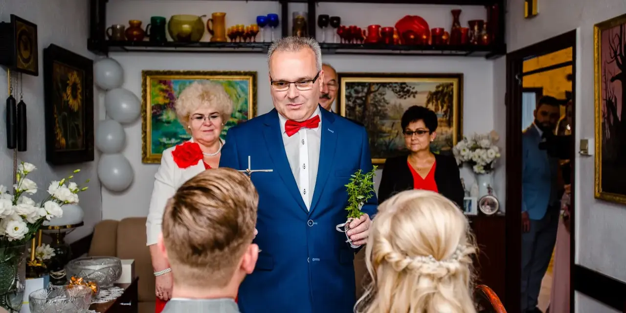 Mężczyzna w niebieskiej marynarce dokonuje tradycyjne błogosławieństwo, trzymając krzyż i gałązkę.