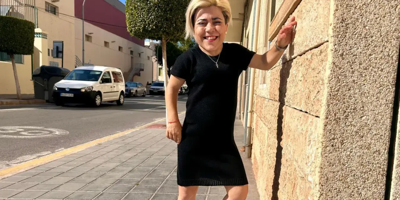 Mujer sonriente con vestido negro, apoyada en pared de piedra. Sus medidas cuerpo mujer son únicas y hermosas.