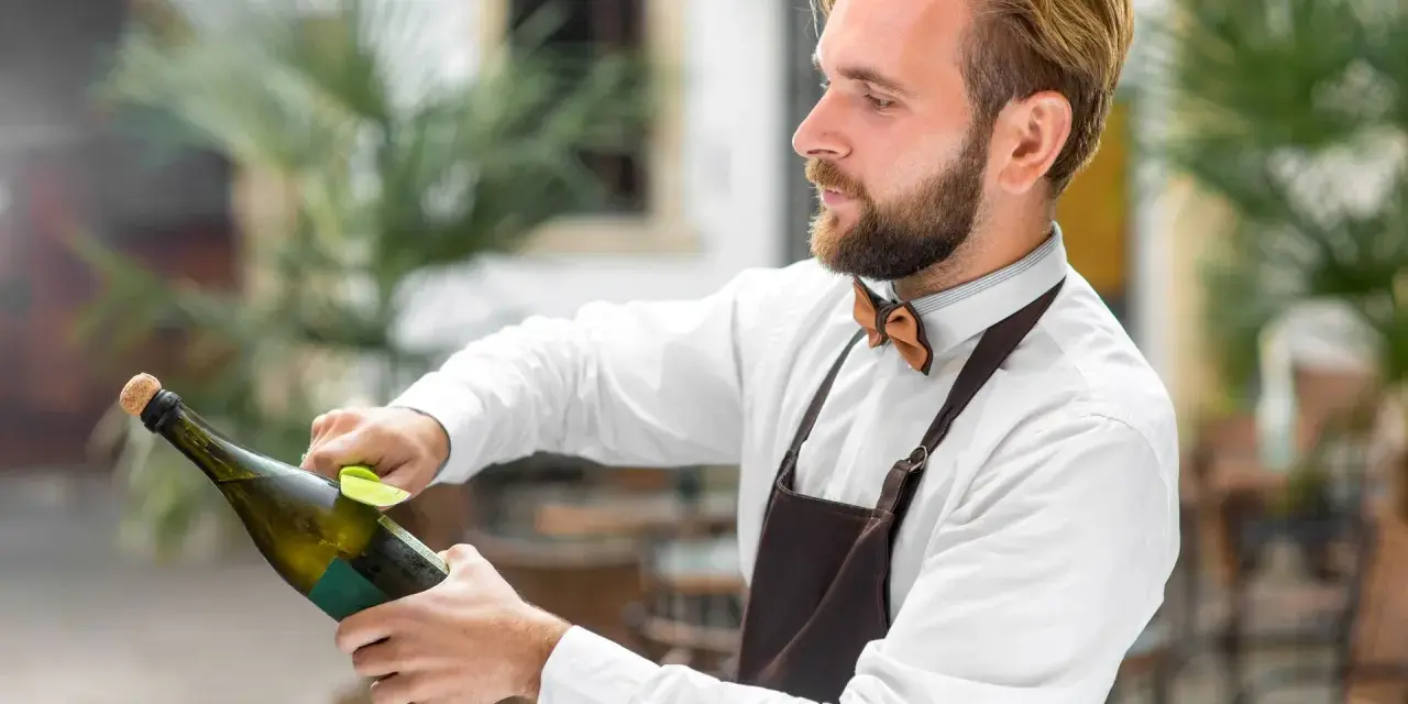 Barman w białej koszuli i brązowym fartuchu pokazuje, jak otwierać szampana, trzymając butelkę i wyciągając korek.
