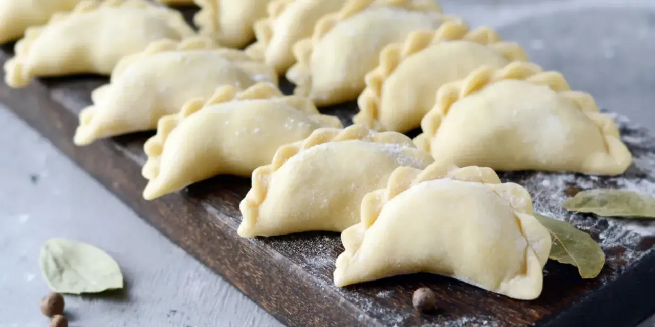 Surowe pierogi ułożone rzędami na drewnianej desce, posypane mąką.