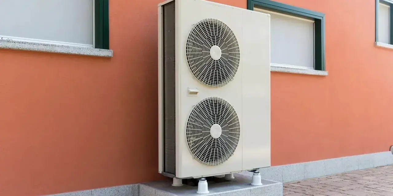 Unità esterna di una pompa di calore, con due ventole, montata su una base rialzata contro un muro arancione con finestre.