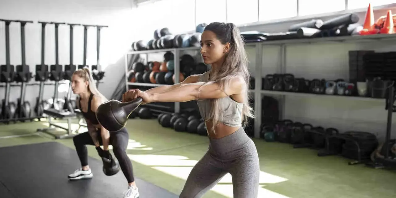 Dwie kobiety ćwiczą z kettlebellami, redukując tkankę tłuszczową. W tle sprzęt treningowy.