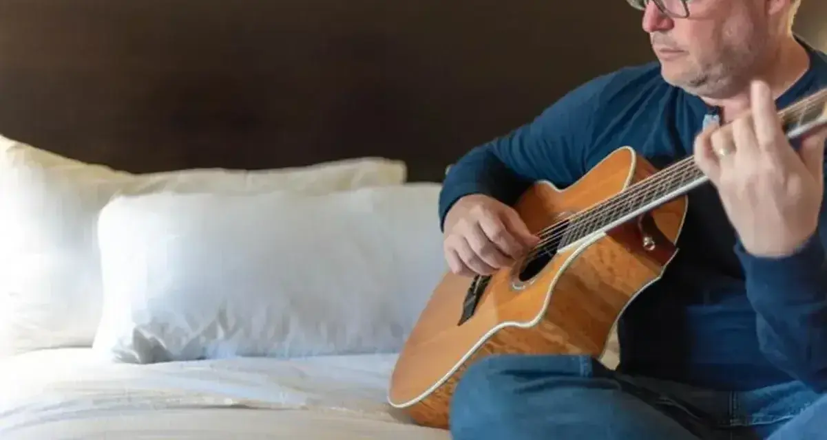 Mężczyzna w okularach gra na gitarze siedząc na łóżku. Gdzie ta keja nuty?