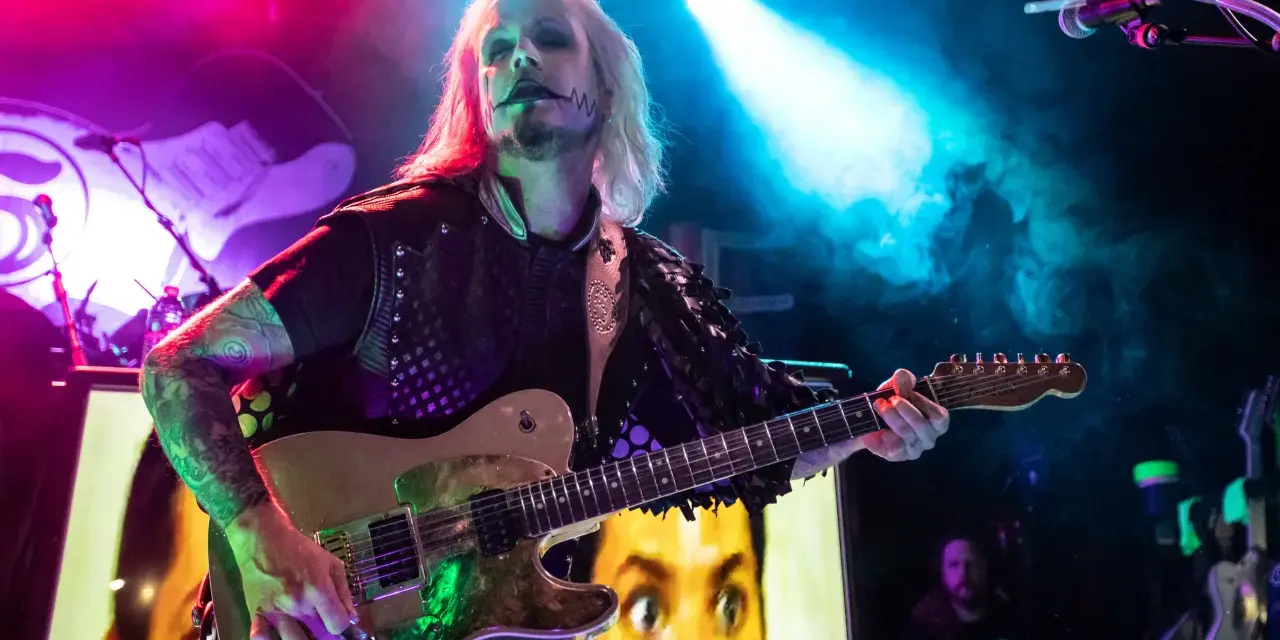 John 5 mit seinem charakteristischen Make-up spielt Gitarre, im Hintergrund ein Bild von Marilyn Manson.