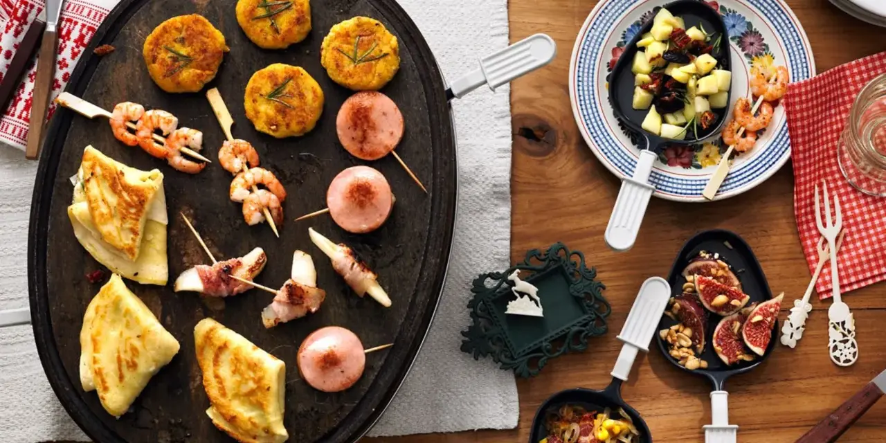 Raclette-Ideen für Kinder: Garnelen-Spieße, kleine Frikadellen, Käse-Speck-Röllchen und Crêpes auf einem Raclette-Grill.