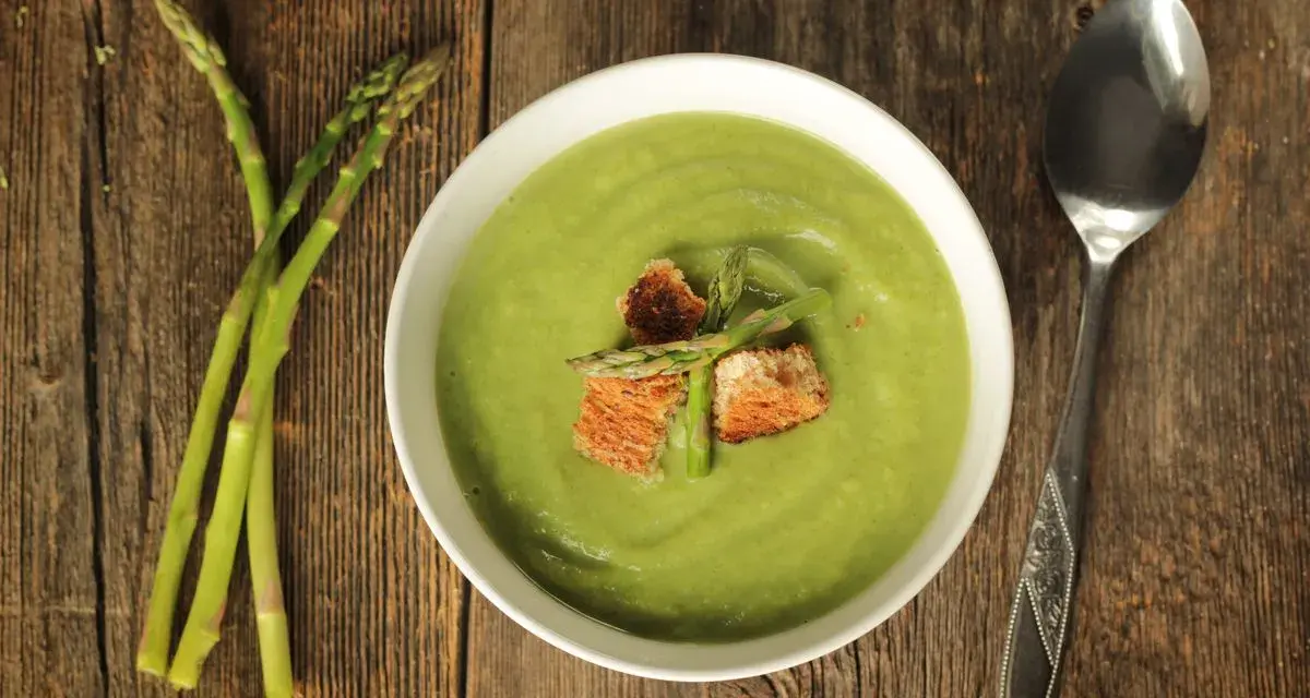 Ciotola di vellutata verde brillante, guarnita con crostini e asparagi freschi. Accanto, un mazzetto di asparagi e un cucchiaio. Un delizioso pesto di asparagi pronto da gustare.