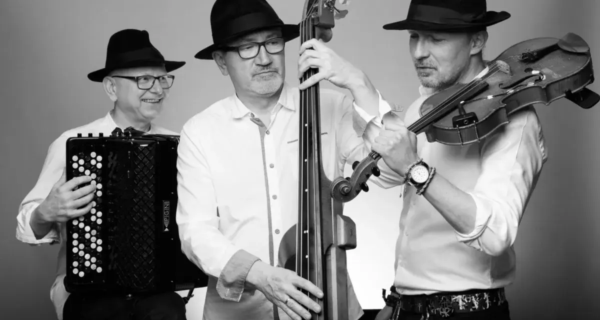 Trzech muzyków w czarnych kapeluszach grających muzykę klezmerską: akordeonista, basista i skrzypek.