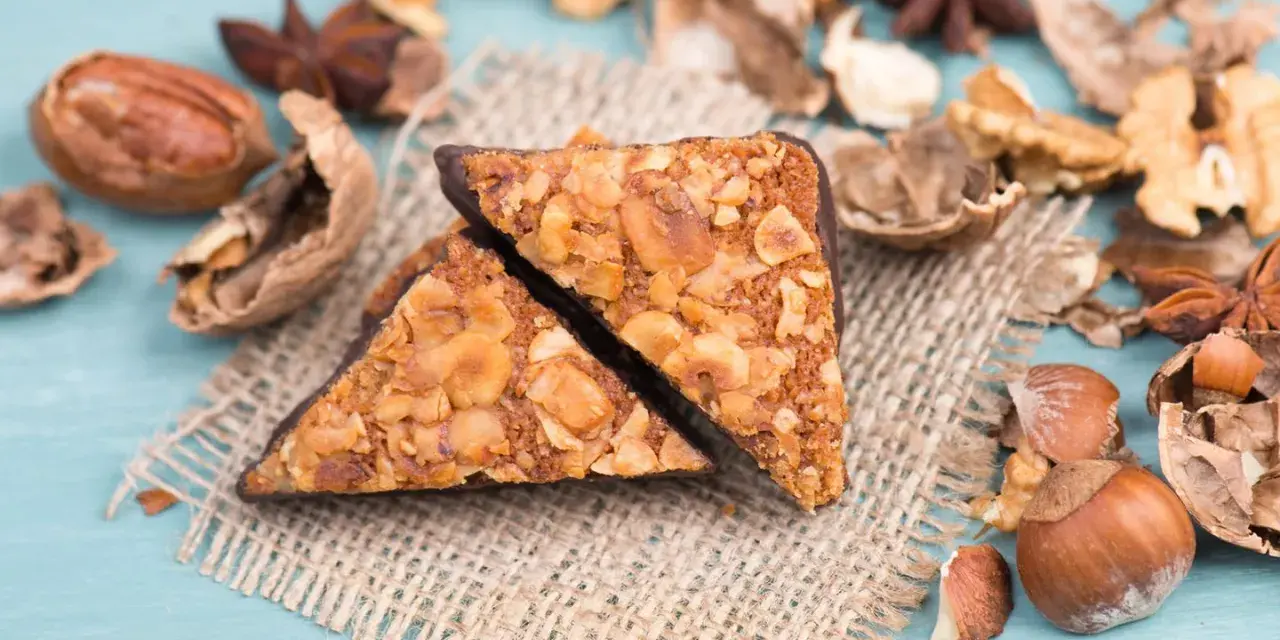 Zwei vegane Nussecken mit Schokoladenrand, bedeckt mit gehackten Nüssen, liegen auf einem Jutesack, umgeben von verschiedenen Nüssen und Sternanis.