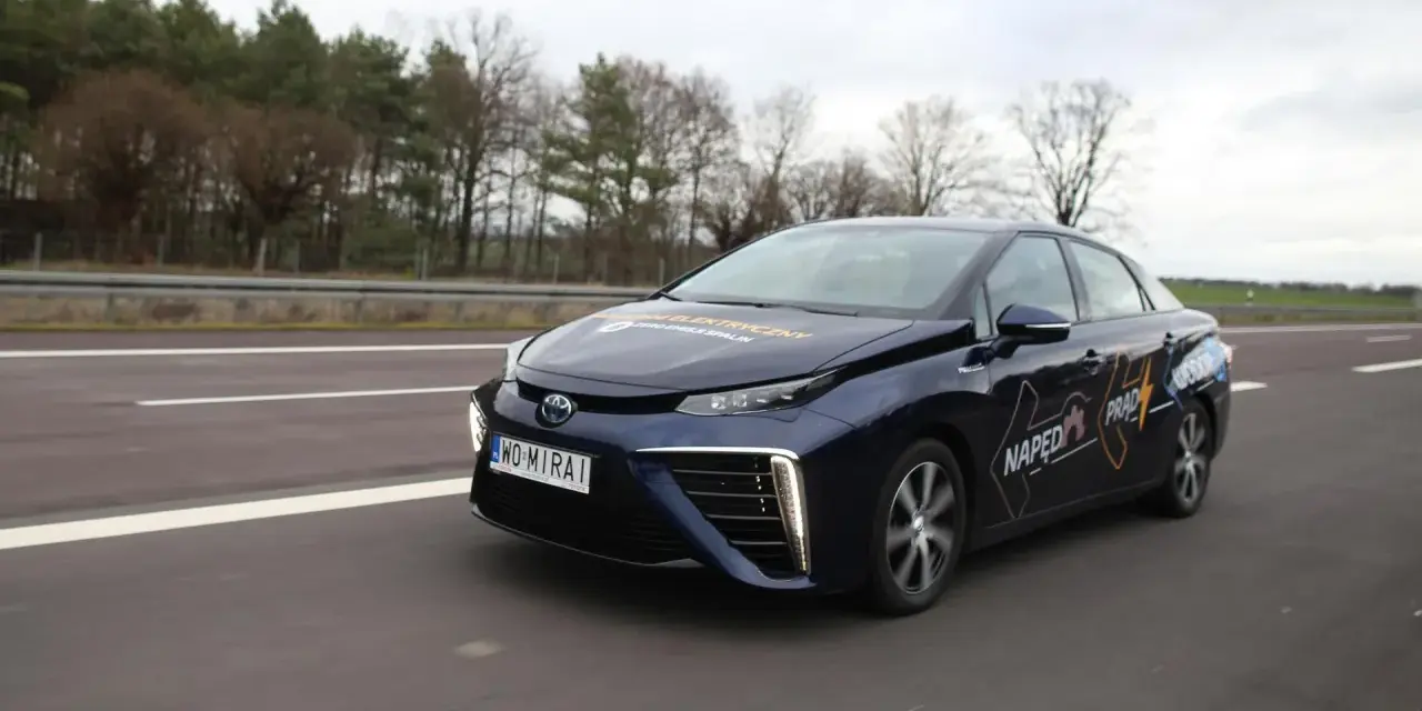 Ciemnoniebieski samochód na wodór Toyota Mirai jedzie po drodze. Na drzwiach widoczny napis "Napęd Prąd Wodór".