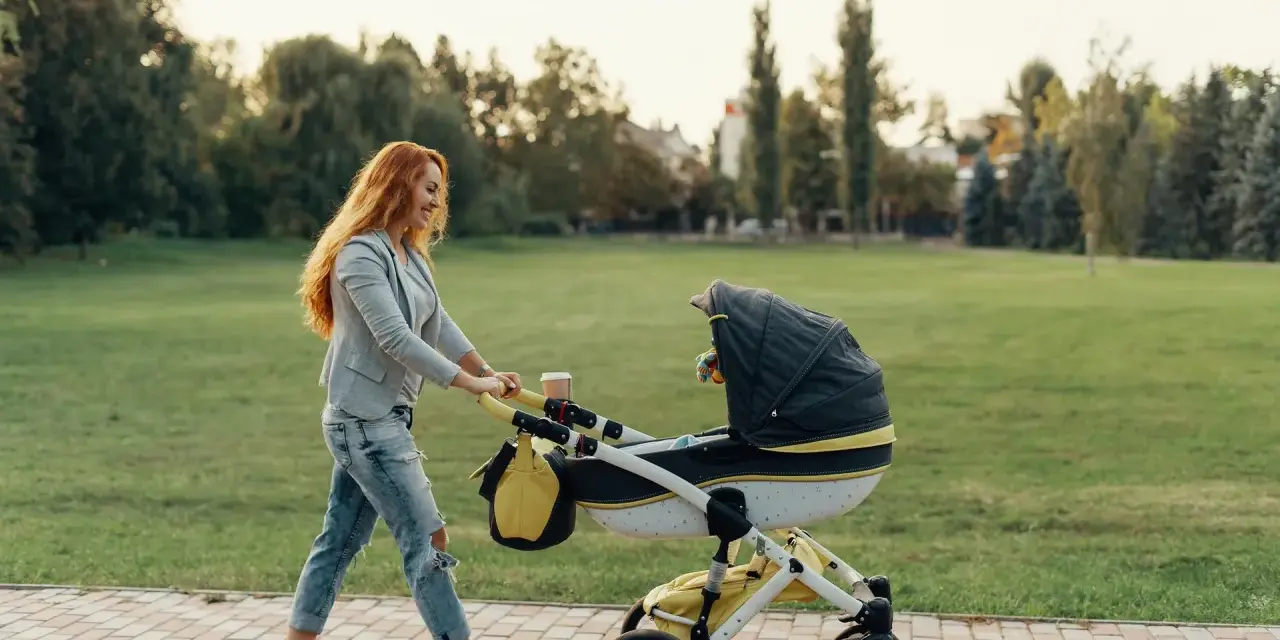 Ruda kobieta z uśmiechem pcha wózek podczas pierwszego spaceru z noworodkiem w parku.