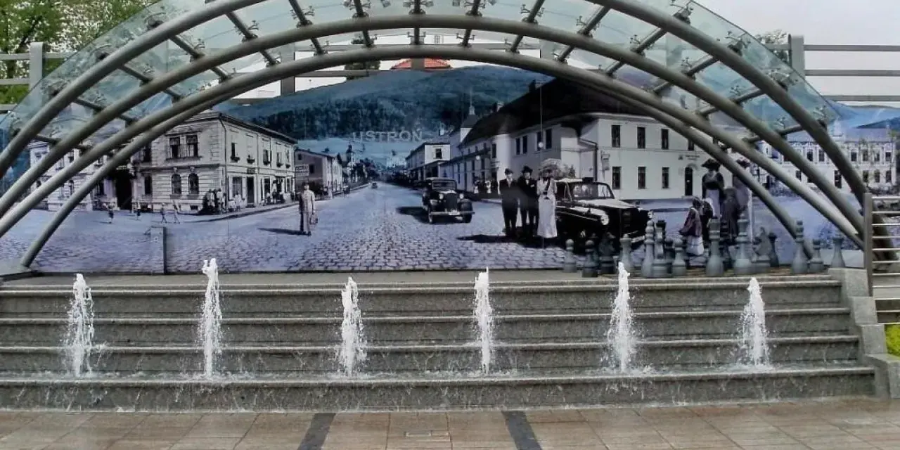 Ustroń rynek: fontanna i mural przedstawiający historyczne miasto z ludźmi w strojach z epoki i zabytkowymi samochodami.