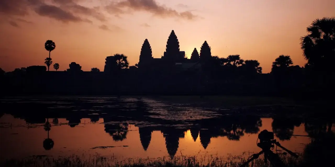 Zachód słońca nad Angkor Wat, idealne tło dla niezapomnianych kambodżańskich wakacji. Widok odbija się w wodzie, a na pierwszym planie statyw z aparatem.