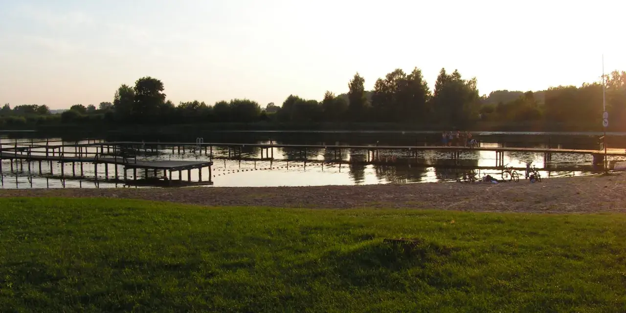 Drewniane pomosty na spokojnym jeziorze starogrodzkim, z piaszczystą plażą i zieloną trawą w tle.