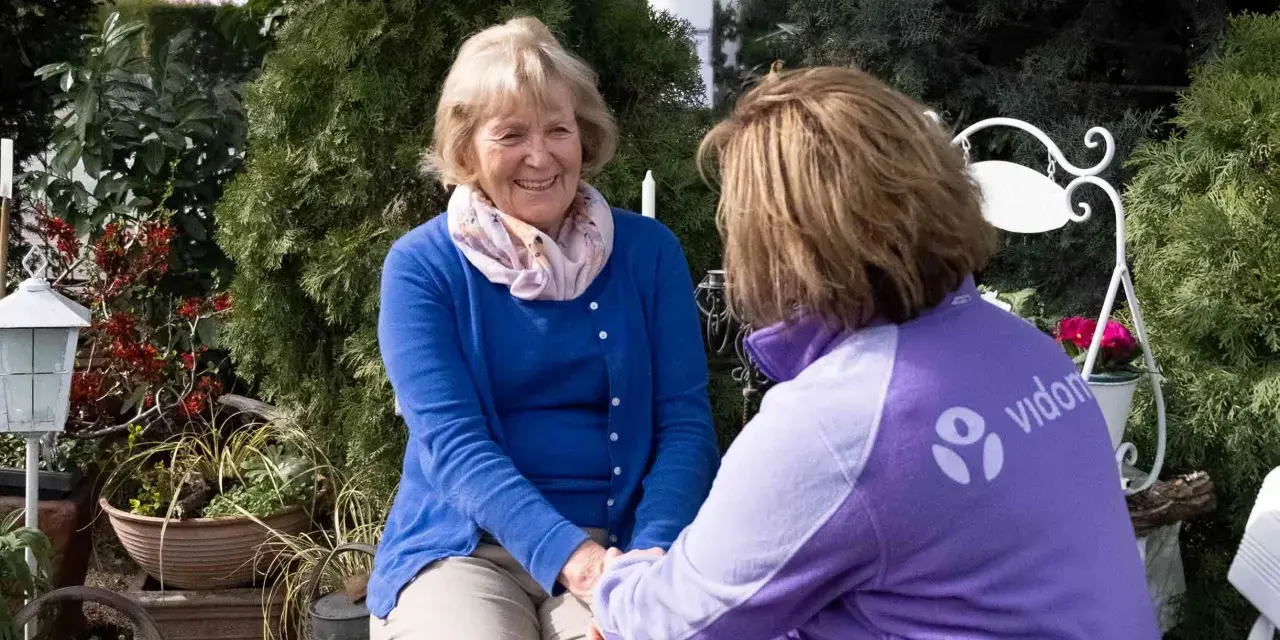 Eine freundliche ältere Dame im Gespräch mit einer Pflegekraft. Deren Kleidung trägt das Logo "vidon". Ein Gutachter für den Pflegegrad könnte hier im Gespräch sein.