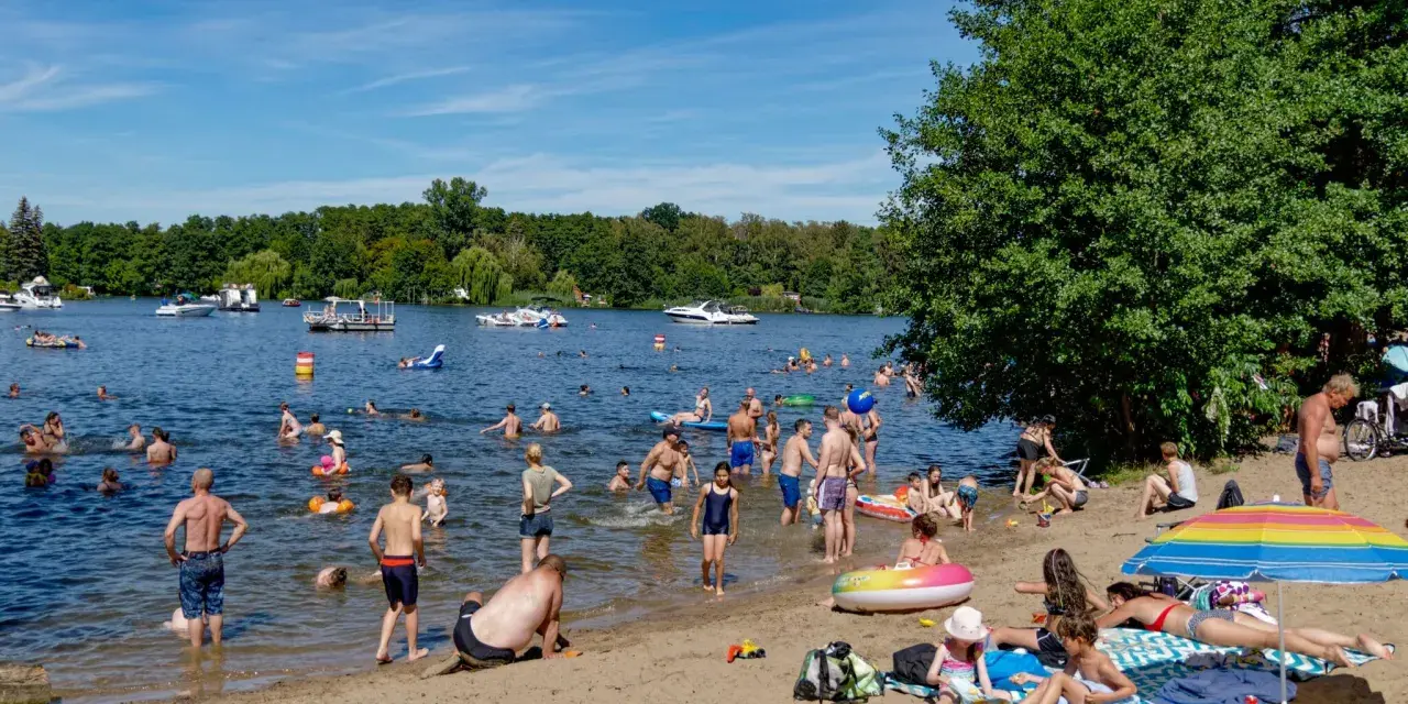 Menschen genießen das Reschensee baden an einem sonnigen Tag. Kinder planschen, Erwachsene schwimmen und entspannen am Ufer. Boote gleiten über das Wasser.