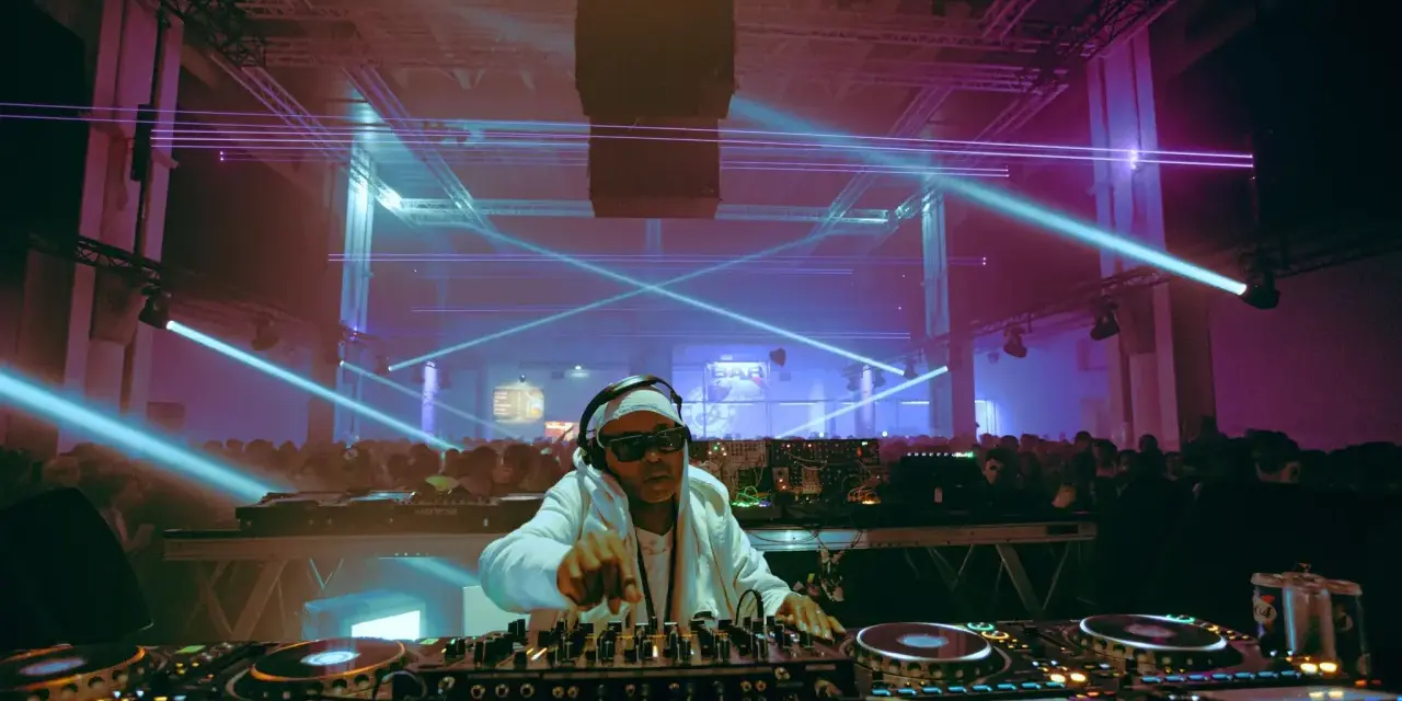 Un DJ année 2000, lunettes de soleil et bandeau, mixe sous des lasers bleus et violets devant une foule en délire.
