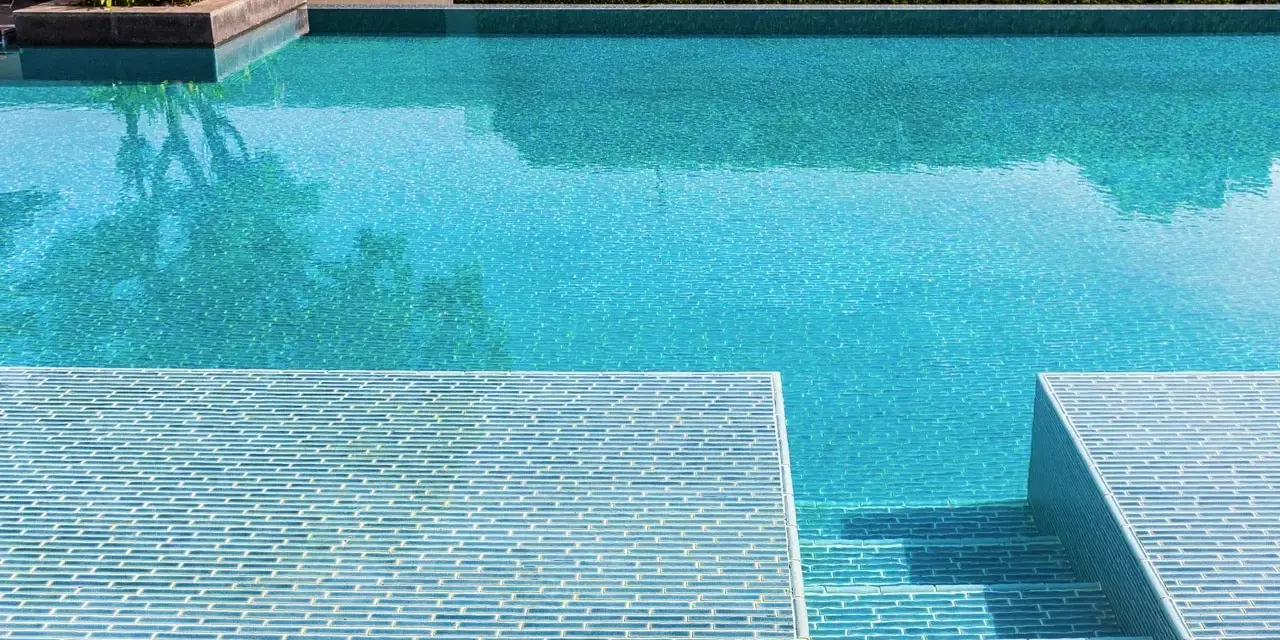 Élégante rénovation piscine mosaïque bleue, avec des marches carrelées et une végétation luxuriante.