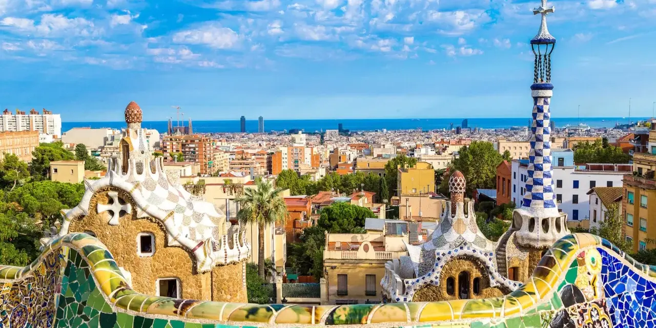 Kolorowe mozaiki i fantazyjne budowle Parku Güell, dzieło Antonio Gaudiego, na tle błękitnego nieba i Barcelony.
