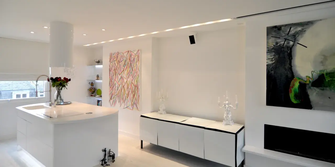 Modernt kök och vardagsrum med vita ytor, konst och spotlights som skapar en ljus och inbjudande atmosfär.