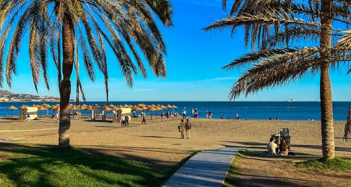 Słoneczna plaża w Maladze z palmami, parasolami i ludźmi spacerującymi brzegiem morza.