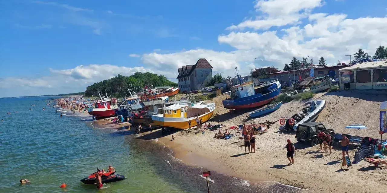 Słoneczny dzień nad morzem, plaża pełna ludzi, kolorowe łodzie na piasku. W tle widać budynki, a na wodzie pływa skuter. Idealne miejsce na wakacje, jakby ktoś przyjechał tu autobusem nad morze.