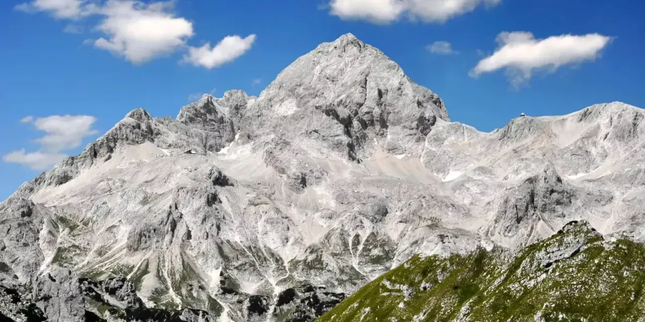 Majestätischer Triglav im Sonnenschein, bereit für die Triglav-Besteigung. Grüne Hänge und Felsen unter blauem Himmel mit Wolken.